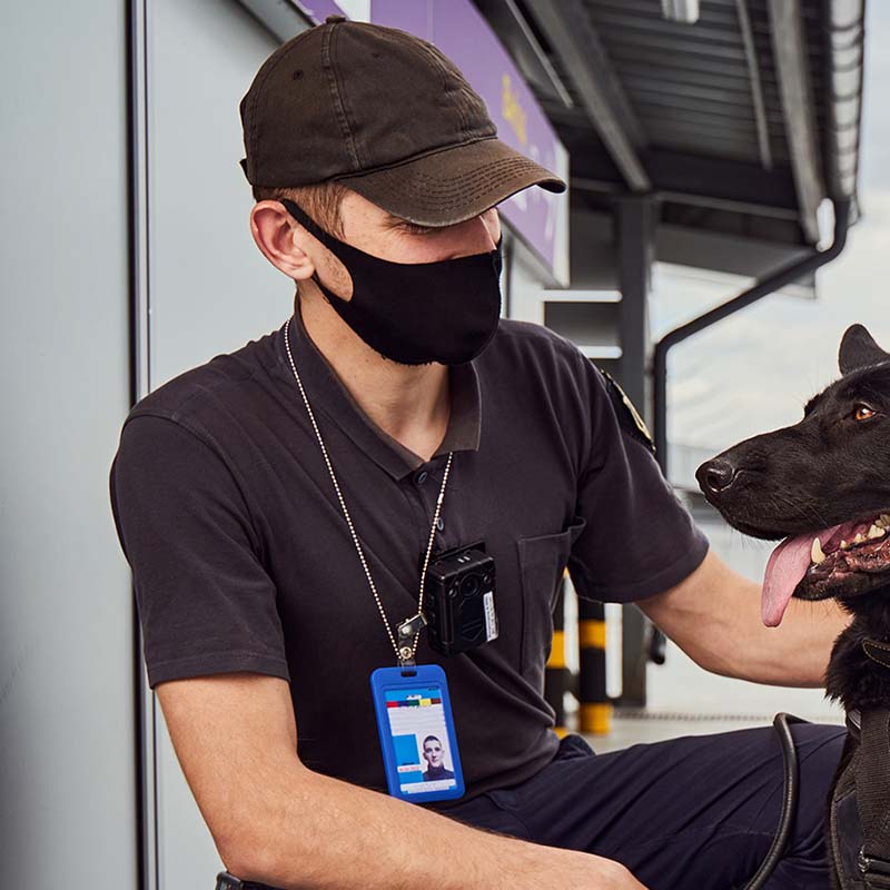 Young man security worker wearing protective face mask while squatting down and looking at black police dog