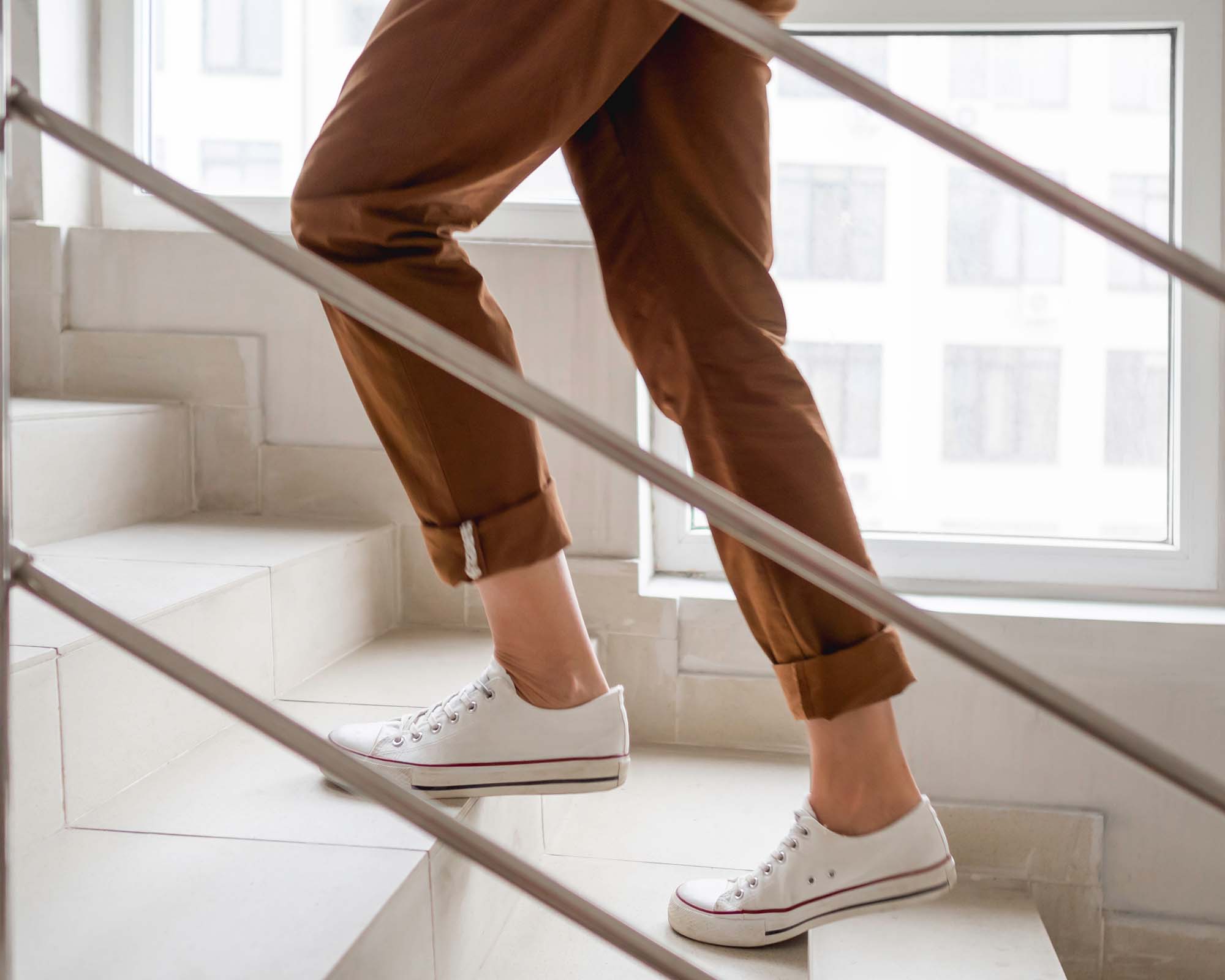 Woman in white sneakers and khaki trousers goes upstairs to her apartment. White staircase in apartment building. Casual outfit, urban fashion.