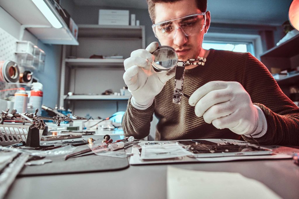 Electronic technician mending a broken tablet carefully examines the chip of the tablet using a magnifying glass in a repair shop.