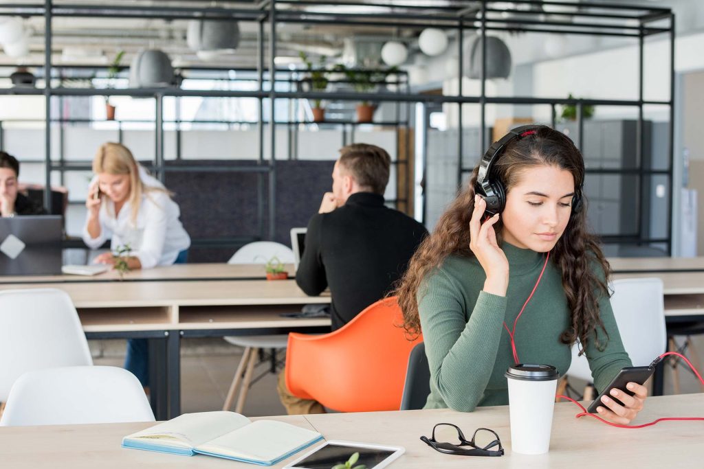 Portrait of contemporary young woman listening to music at workplace wearing big headphones in open space office