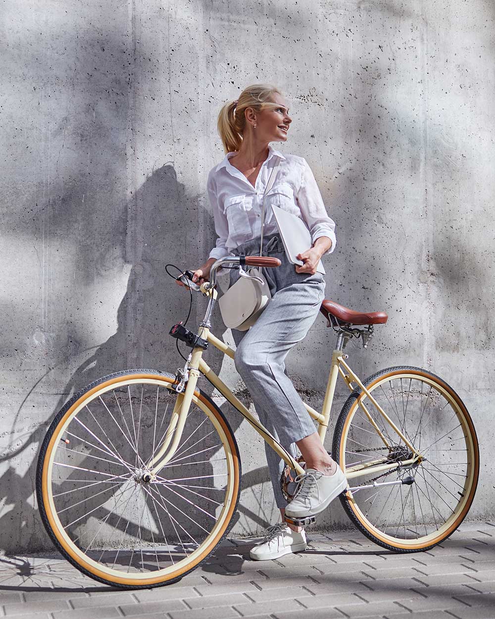 Full length portrait of fashionable woman in business casual clothes holding her hands on handlebar of a city bike isolated on the grey wall
