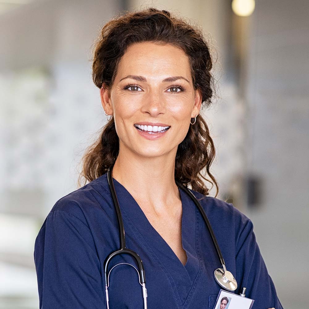 Portrait of happy young female nurse with folded arms standing in hospital hallway. Confident doctor woman in uniform and stethoscope looking at camera with copy space. Portrait of beautiful young healthcare worker working in private clinic.