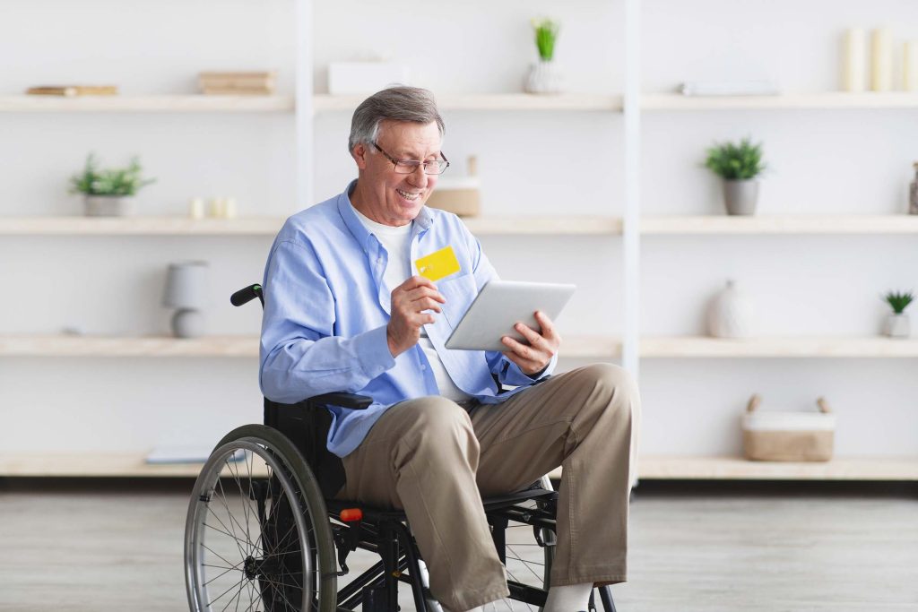 Happy senior man in wheelchair shopping online with tablet computer and credit card at home. Positive older male with disability buying things at internet store, using electronic money