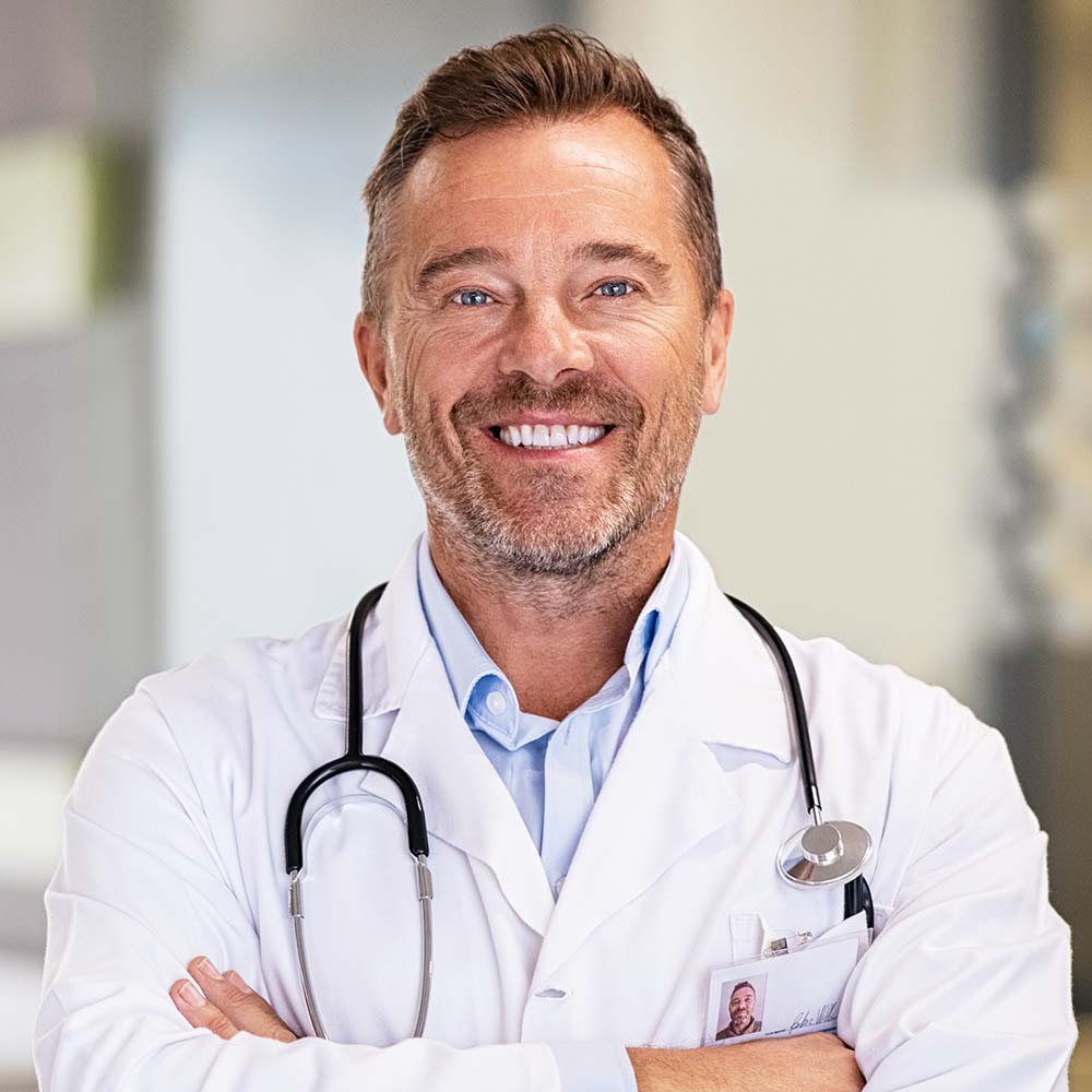 Portrait of happy mature doctor with folded arms standing at hospital hallway. Confident male doctor in a labcoat and stethoscope looking at camera with satisfaction. Smiling confident general practitioner at private health clinic.