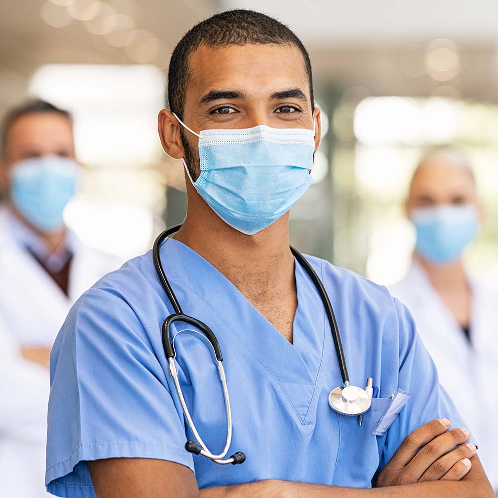 Confident multiethnic male nurse in front of his medical team looking at camera wearing face mask during covid-19 outbreak. Happy and proud indian young surgeon standing in front of his colleagues wearing surgical mask for prevention against coronavirus. Portrait of mixed race doctor with medical staff in background at hospital.