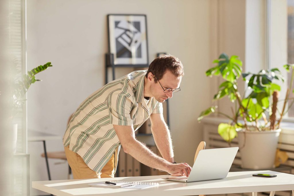 Horizontal medium shot of modern Caucasian man wearing eyeglasses standing at desk in modern office checking something on laptop