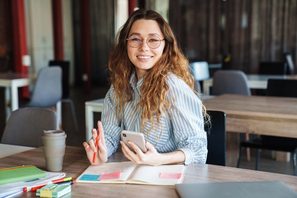 Image of joyful charming woman using cellphone and smiling while studying at classroom