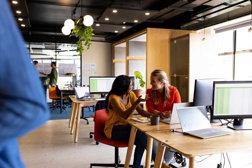 Diverse female colleagues in discussion using laptop at desk in office. Casual office, teamwork, business and work, unaltered.